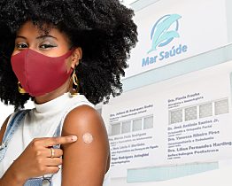 Pessoa mostrando o braço após a vacina, com o painel de vacinas da clínica ao fundo
